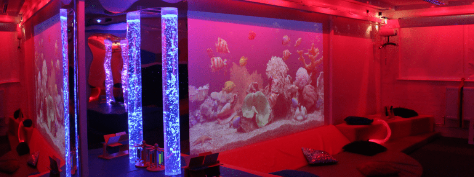 A sensory room bathed in soft red lighting featuring bubble tubes glowing with blue and purple hues, a projected underwater scene with tropical fish on the wall, and cushioned seating areas with pillows for relaxation.