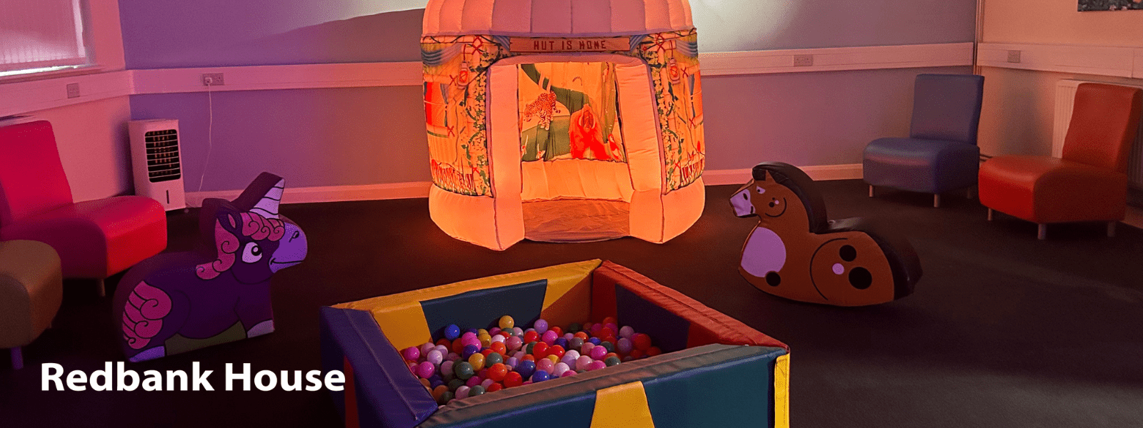 A warmly lit sensory room at Redbank House featuring a soft play area with a colourful ball pit, inflatable hut labeled "HUT IS HOME", padded ride-on toys shaped like a unicorn and a horse, and brightly coloured chairs arranged around the edges of the room.